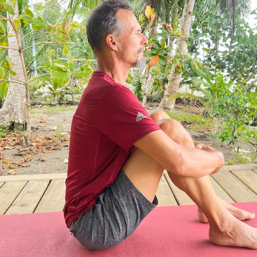A man sits cross-legged on a yoga mat and meditates outdoors under trees.