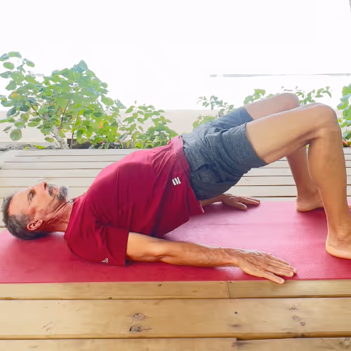 A man does a bridge exercise on a yoga mat outdoors with plants in the background.