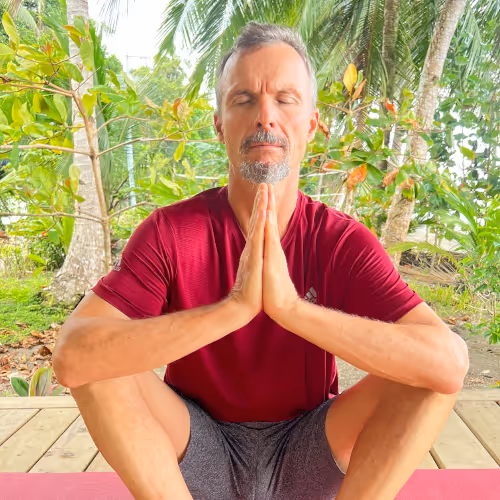 A man in a red T-shirt and grey shorts meditates outdoors with closed eyes and folded hands in front of tropical plants.