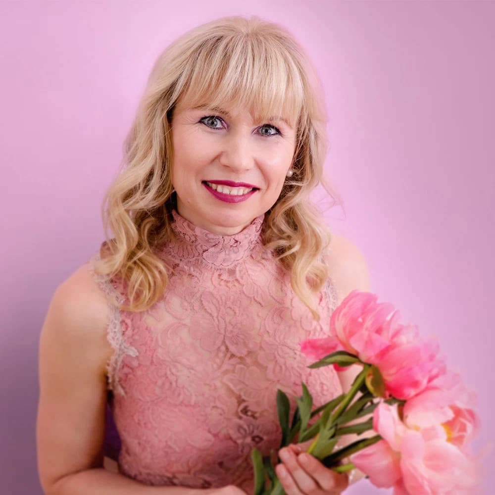 A woman with blonde curly hair in a pink lace dress holds a bouquet of pink flowers in front of a pink background, this is Veronika Mutti, expert in massage and coaching in Switzerland