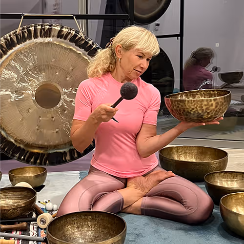 A woman in lotus position holds a singing bowl and strikes it with a mallet, surrounded by several singing bowls and a gong.