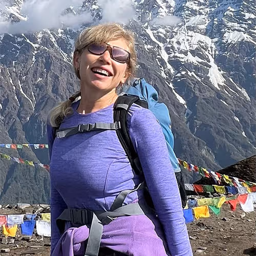 A woman with sunglasses and a rucksack, laughing in front of snow-covered mountains and colourful prayer flags.