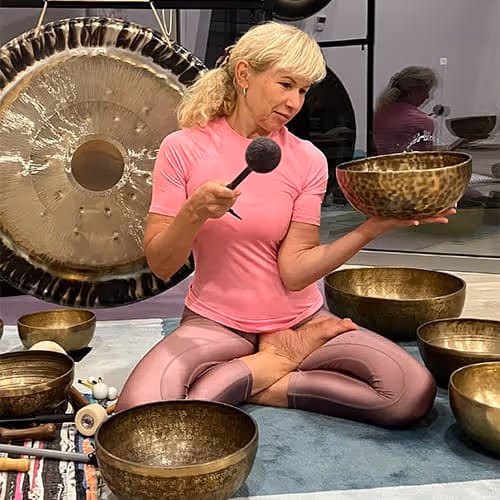 A woman in a yoga seat with singing bowls in a meditation room, holding a bowl and a mallet.