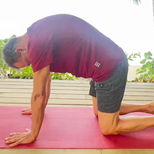 A man in a red T-shirt and grey shorts does the cat stretch exercise on a red mat, looking down.