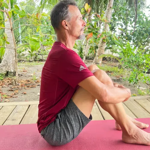 A man sits cross-legged on a yoga mat and meditates outdoors under trees.