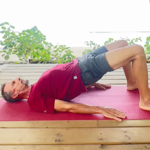 A man does a bridge exercise on a yoga mat outdoors with plants in the background.