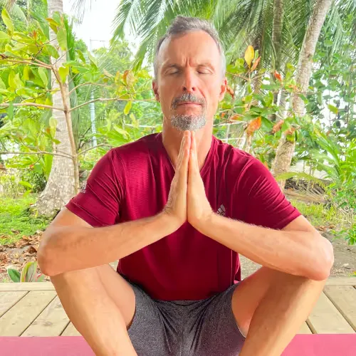 A man in a red T-shirt and grey shorts meditates outdoors with closed eyes and folded hands in front of tropical plants.
