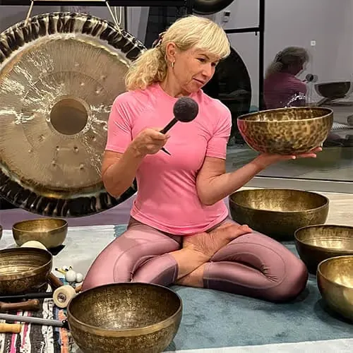 A woman in a yoga seat with singing bowls in a meditation room, holding a bowl and a mallet.