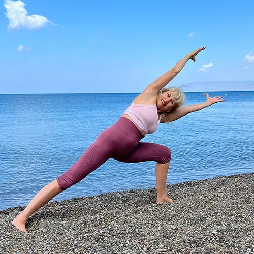 A woman does a yoga stretch on a pebbly beach in front of a calm lake under a blue sky.