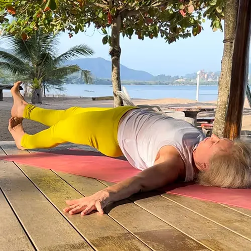 A woman in yellow leggings does a yoga exercise lying on her side on a wooden terrace overlooking beach and sea.