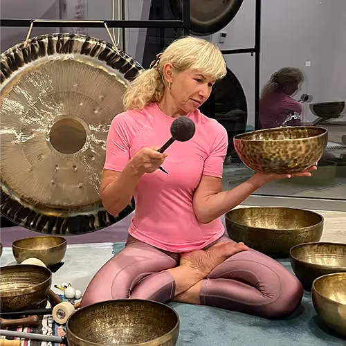 A woman in lotus position holds a singing bowl and strikes it with a mallet, surrounded by several singing bowls and a gong.
